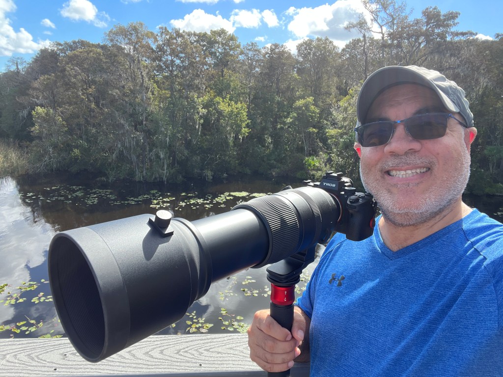 Luis posing with his camera and a long lens resting on a monopod. A lake and woods appear in the background.